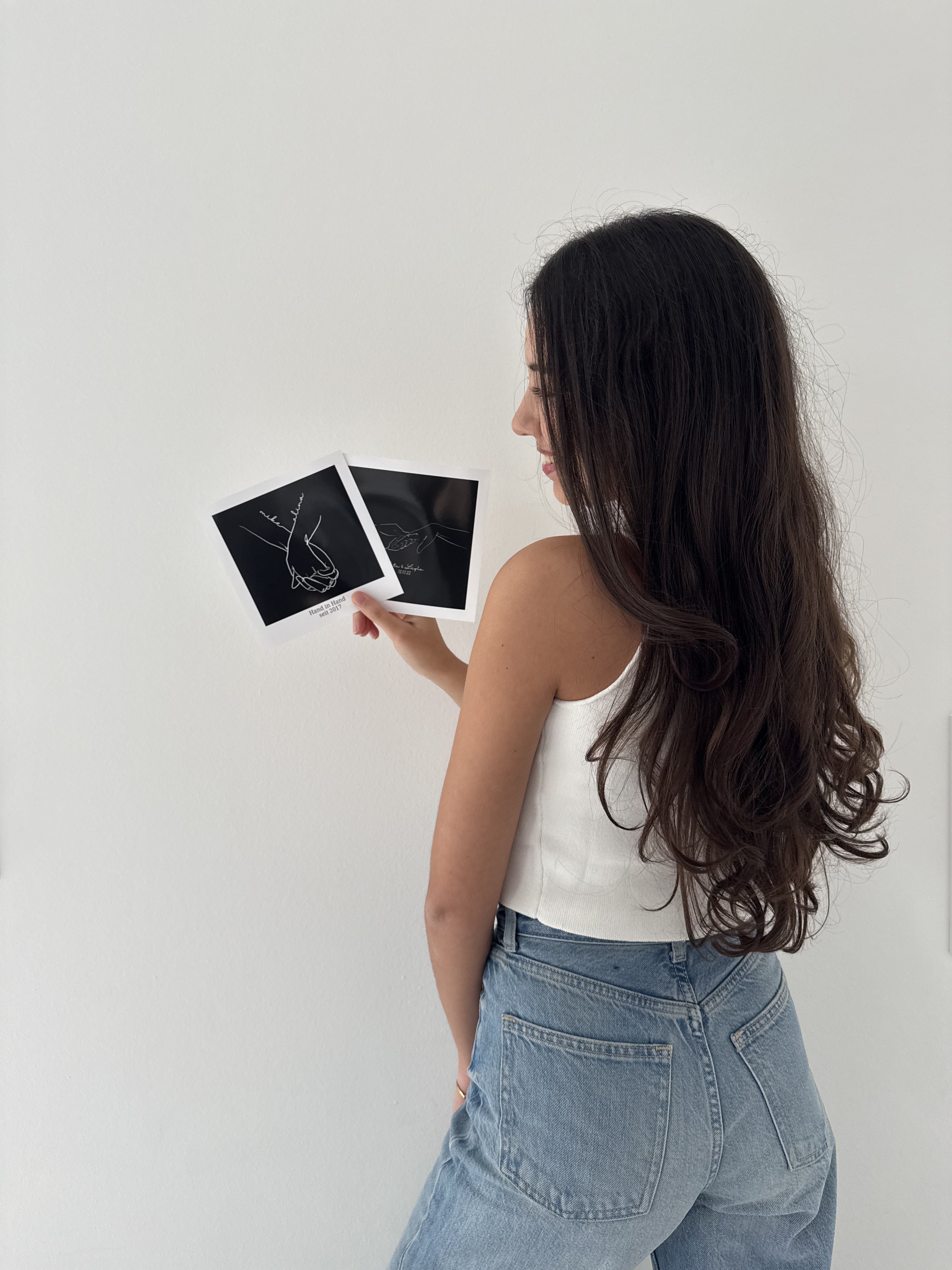 Selin, Gründerin von Sels Studio, steht seitlich vor einer weißen Wand und hält zwei personalisierte Line-Art-Poster in Schwarz in der Hand. Die minimalistischen Illustrationen zeigen Händchen haltende Paare – ein Symbol für Nähe, Liebe und Individualität.