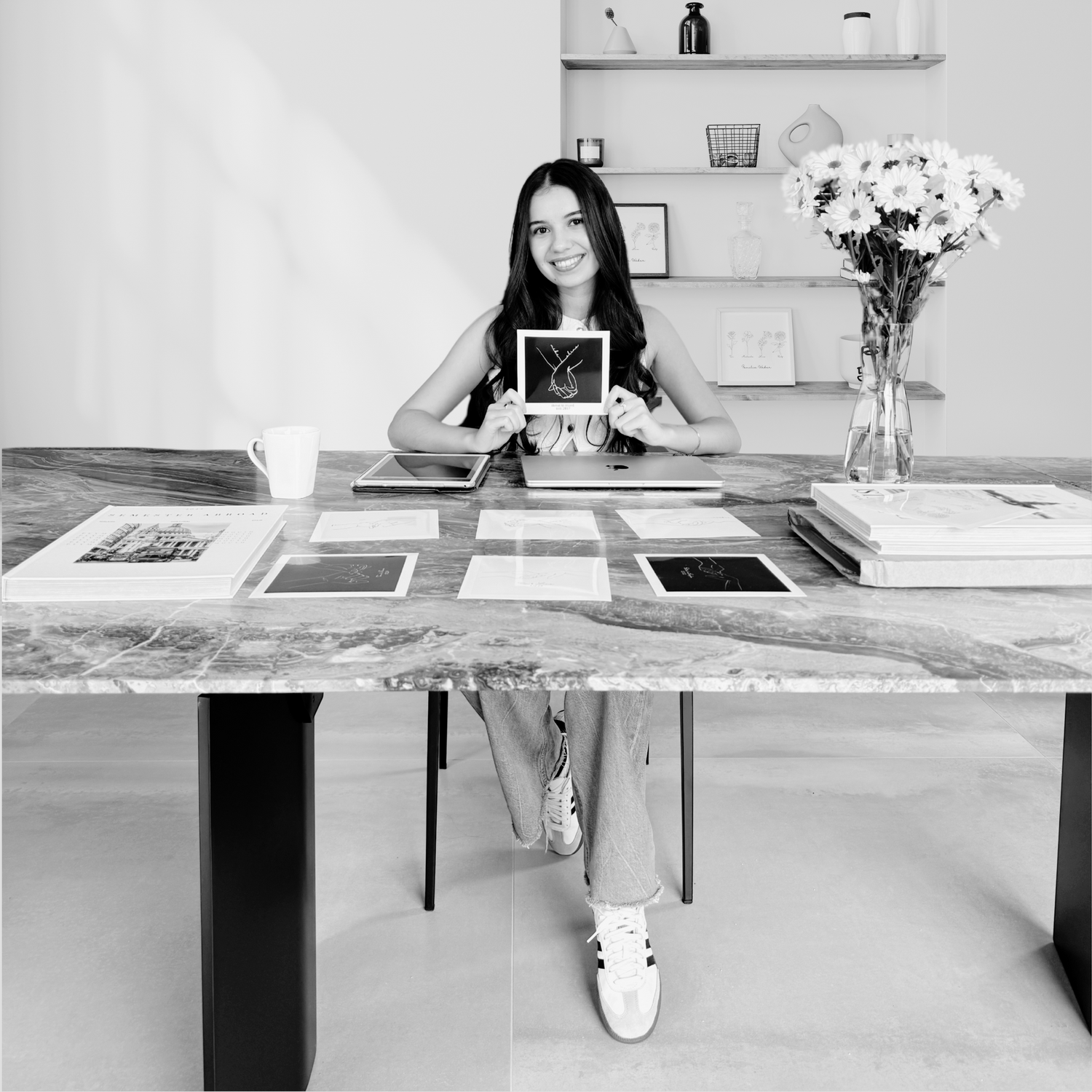 Selin, die Gründerin von Sels Studio, sitzt an einem Steintisch und hält stolz ein „Cheri me Cheri“-Poster in der Farbe Schwarz. Vor ihr liegen weitere personalisierbare Line-Art-Designs ausgebreitet. Im Hintergrund ein Regal mit gerahmten Illustrationen – ein Einblick in die kreative Arbeit und das Herz hinter Sels Studio.
