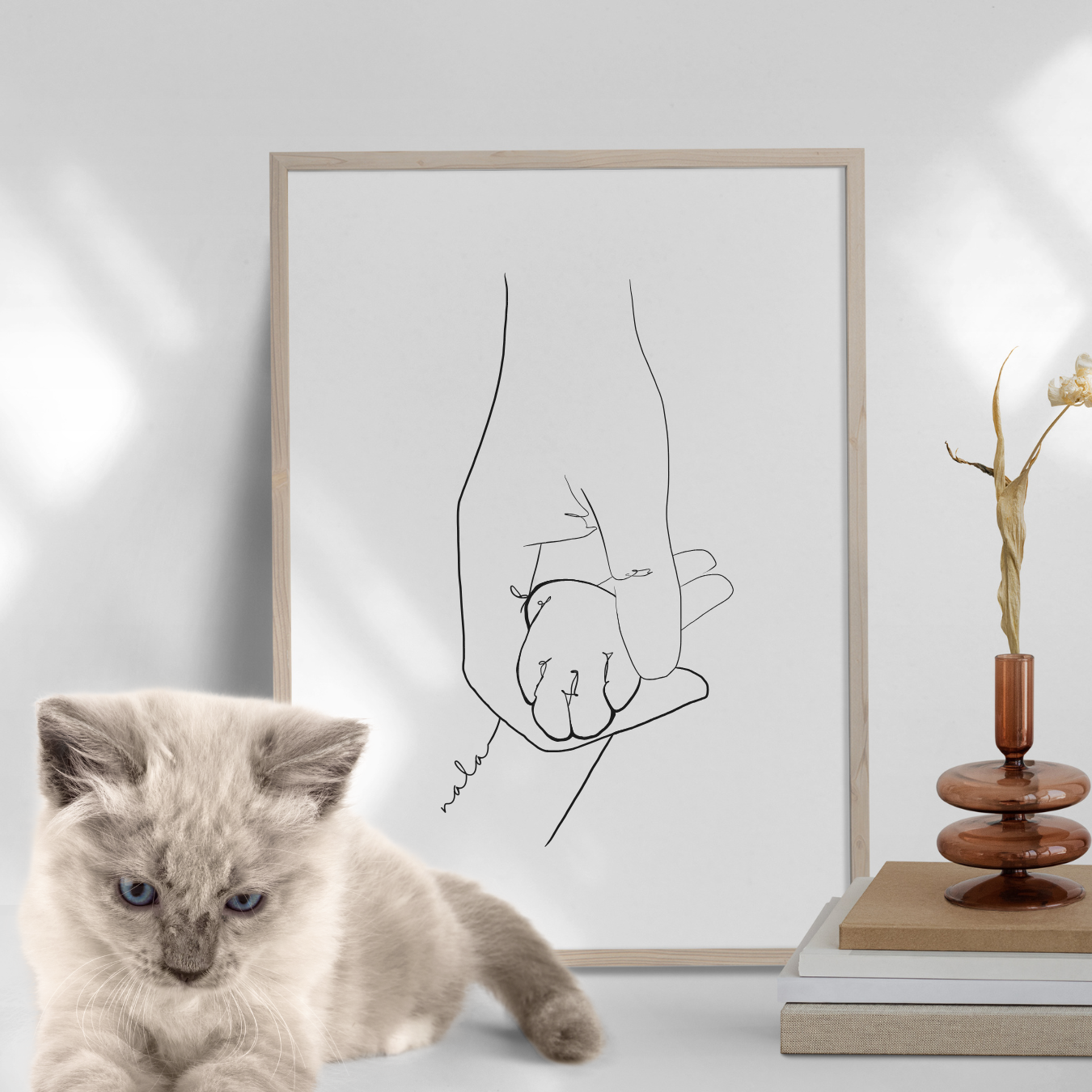 Das Poster 'Pati' zeigt eine stilisierte Hand, die eine kleine Katzenpfote festhält, in minimalistischer Line-Art. Das Kunstwerk ist in einem schlichten Holzrahmen eingebettet und ergänzt mit einer sanften, modernen Atmosphäre, die durch das Kätzchen im Vordergrund noch verstärkt wird.
