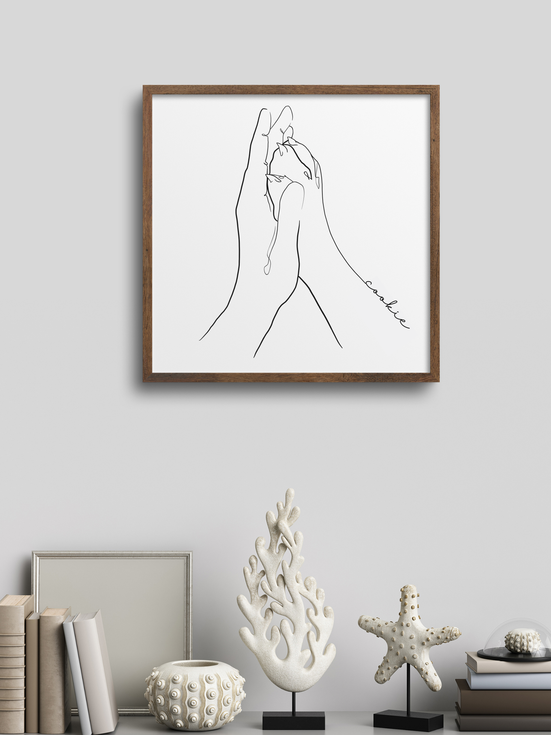 Das Poster 'Patita' zeigt eine stilisierte Hand, die eine kleine Pfote hält, in minimalistischer Line-Art. Das Design ist in einem eleganten Holzrahmen präsentiert und ergänzt durch eine ruhige, moderne Dekoration mit weißen Elementen wie einer Muschel, einem Stern und einem Korallenobjekt.